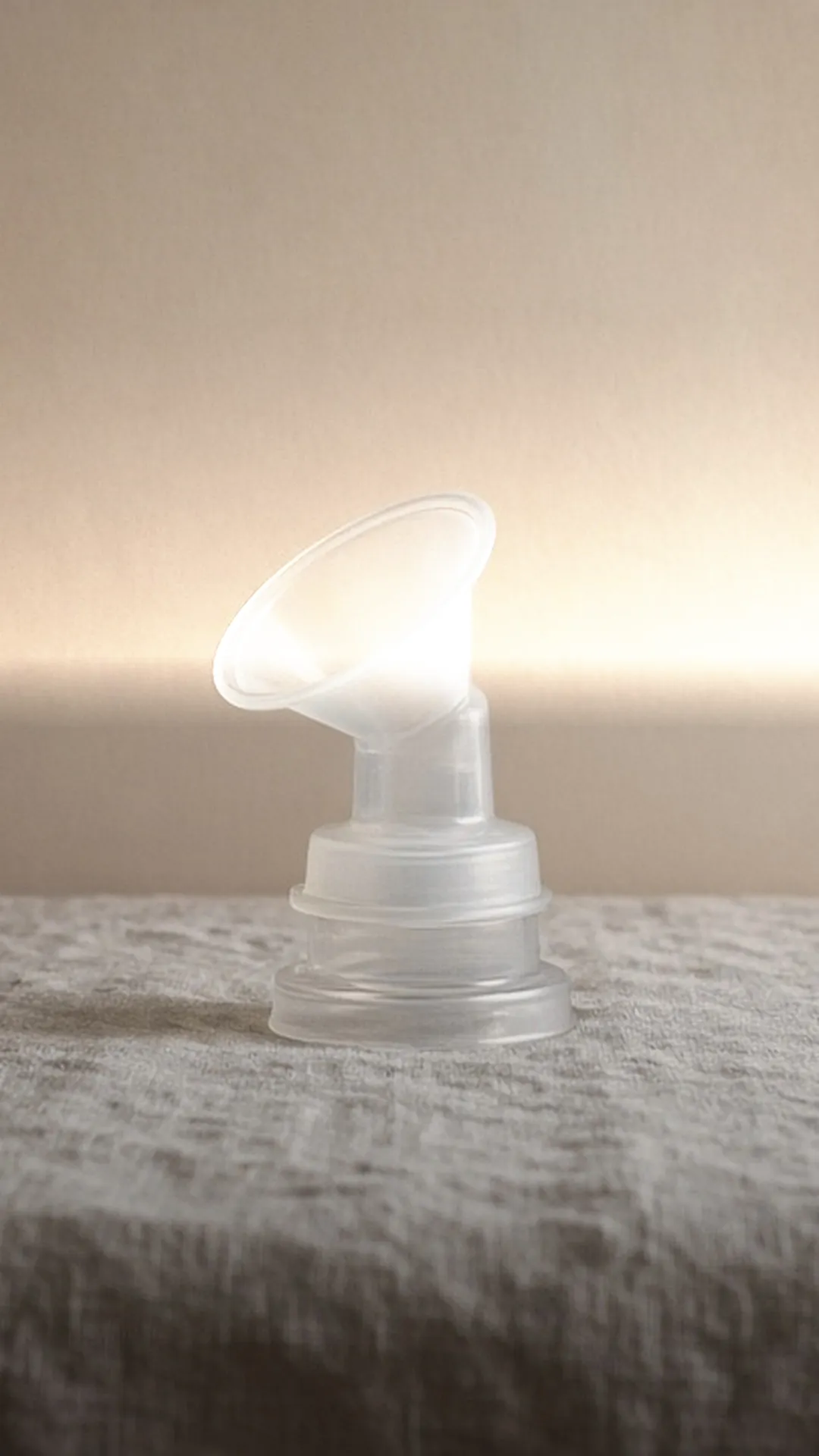One clear breast shield on its connector, standing on grey textured linen